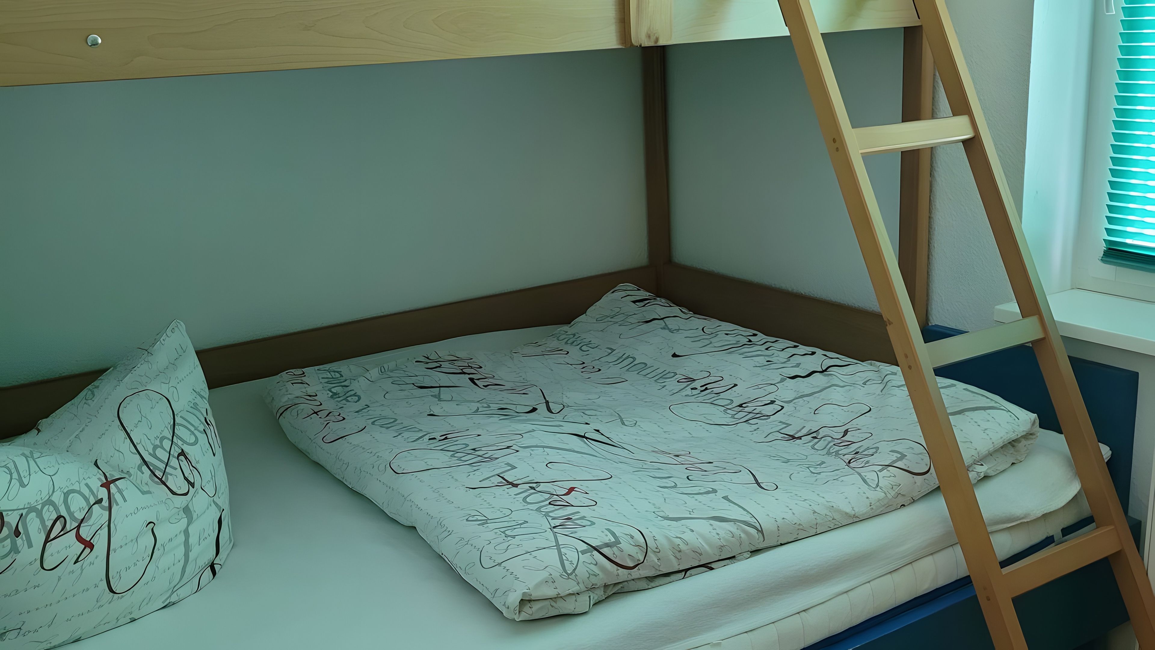 Ein Etagenbett mit einer Leiter, ein Kopfkissen mit Aufdruck und ein Fenster mit Jalousien.