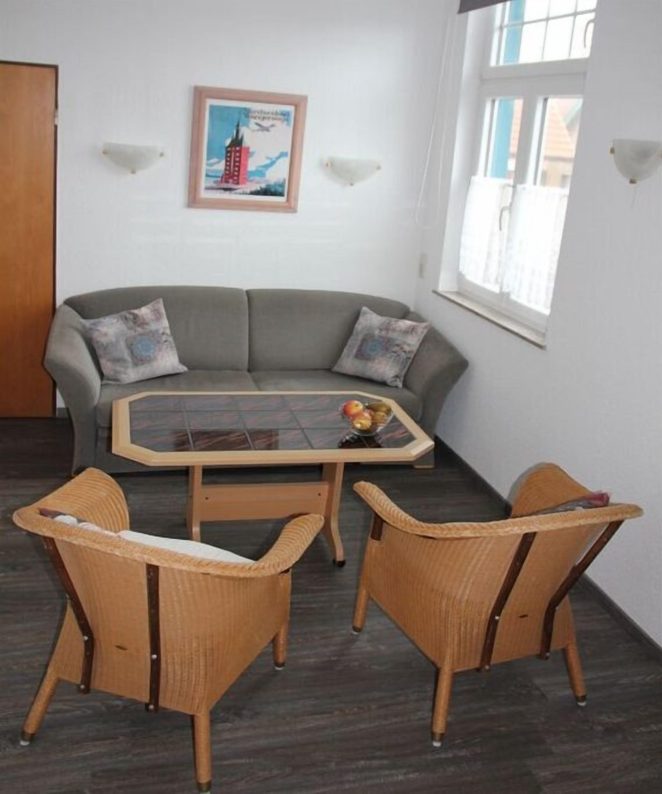 Una habitación con un sofá gris, dos sillas de mimbre, una mesa con superficie de vidrio y fruta, y un cuadro en la pared.