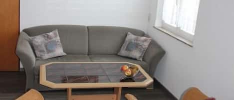 Una habitación con un sofá gris, dos sillas de mimbre, una mesa con superficie de vidrio y fruta, y un cuadro en la pared.