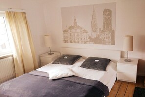A hotel room with a bed, two bedside tables, a lamp, a window with curtains, and a wall-mounted artwork. - Holiday apartment with 1 bedroom (Lüneburg)
