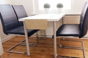 A dining area with a white table and black leather chairs, a radiator, and two potted plants on the windowsill. - Holiday apartment with 1 bedroom (Lüneburg)