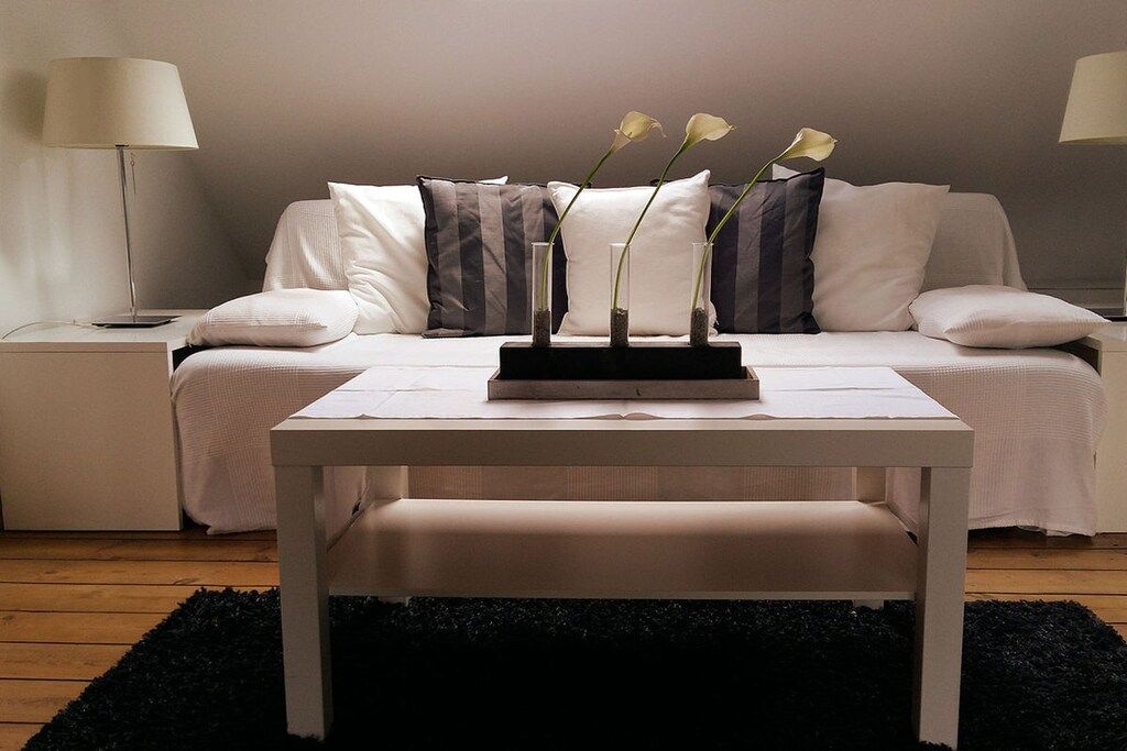 Een slaapkamer met een bed, nachtkastjes, een bijzet-tafeltje, een witte tafel met een bloevas en een zwart vloerkleed.