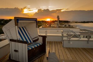A deck with a wicker chair and a cushioned bench, overlooking a body of water with a sunset in the background. - Holiday home with 2 bedrooms (Bitterfeld-Wolfen)