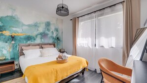 A bedroom with a bed, a chair, a wall painting, and a window with curtains. - Le Maobi · Quiet 2-bedroom apartment with a terrace and parking, 15 minutes from Bordeaux (Tresses, 33370)