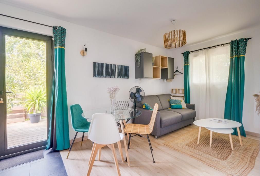 Una sala de estar moderna con una mesa de vidrio, un sofá gris, cortinas verde azulado y vistas a la vegetación.