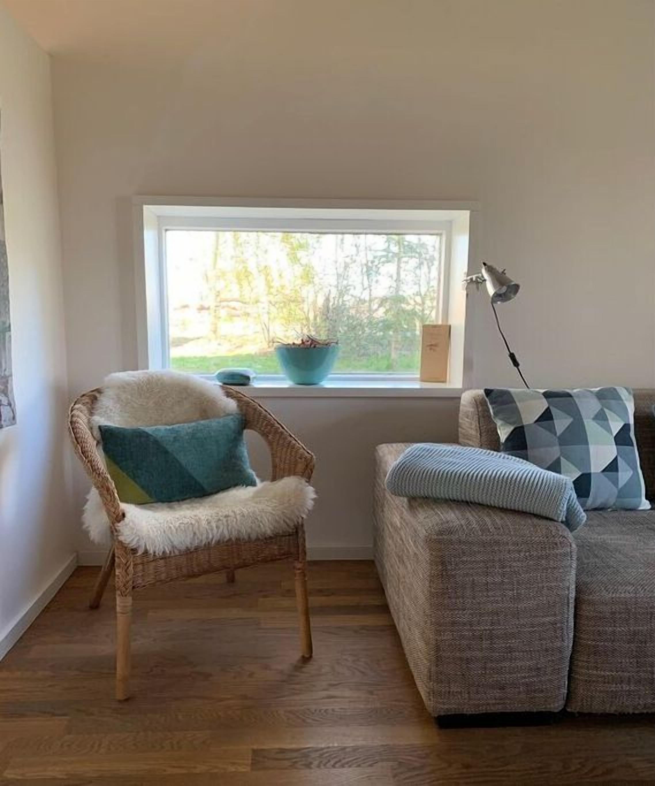 Une pièce avec une fenêtre, un fauteuil en osier avec un coussin, un canapé avec des coussins à motifs et une lampe sur pied.