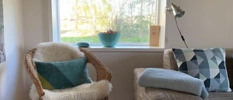 Una habitación con ventana, una silla de mimbre con cojín, un sofá con cojines estampados y una lámpara de pie.