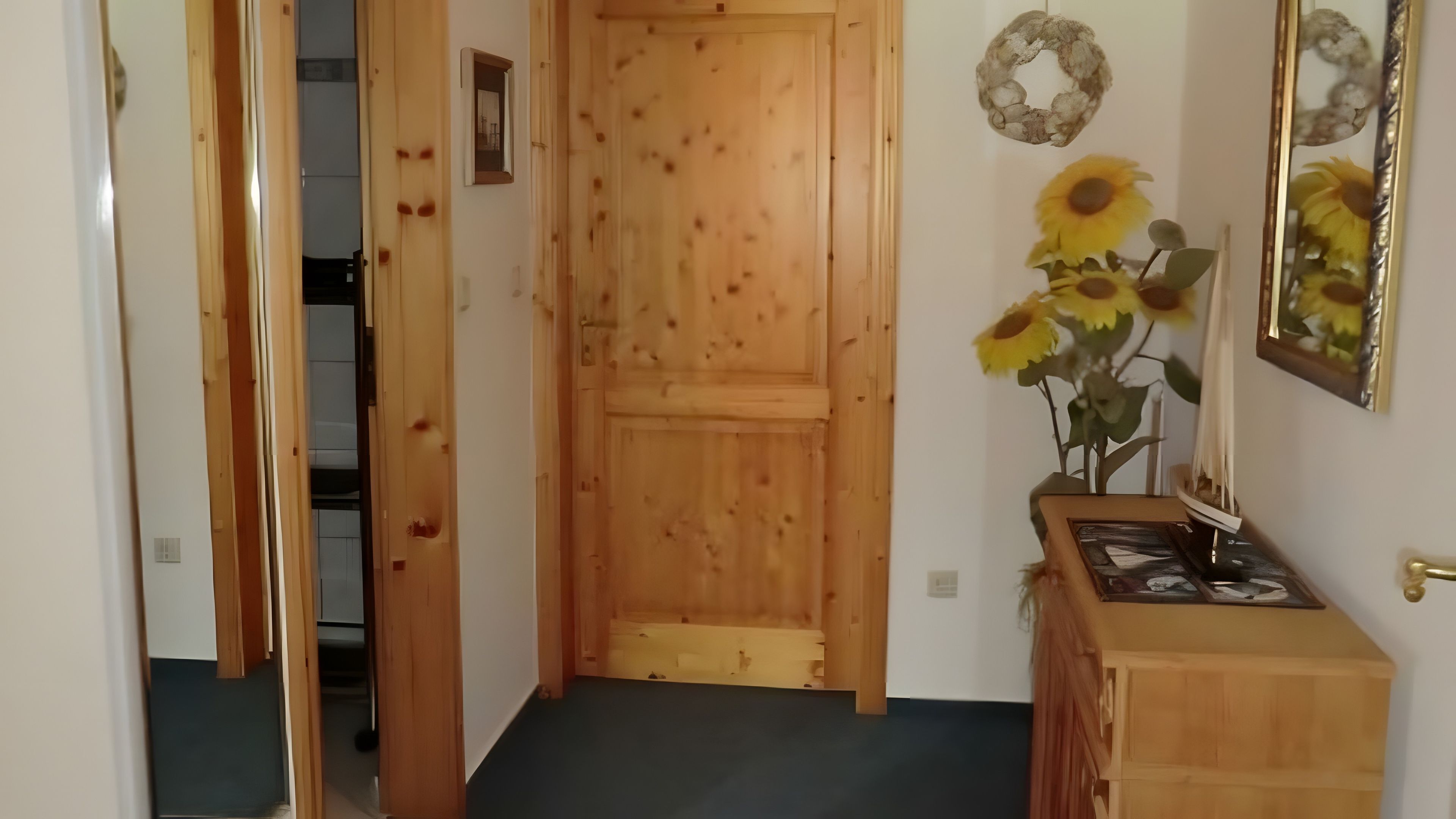 Eine Holztür führt in einen Raum mit dunklem Boden, einem kleinen Holzschrank und einem Wandspiegel mit Sonnenblumenverzierung.