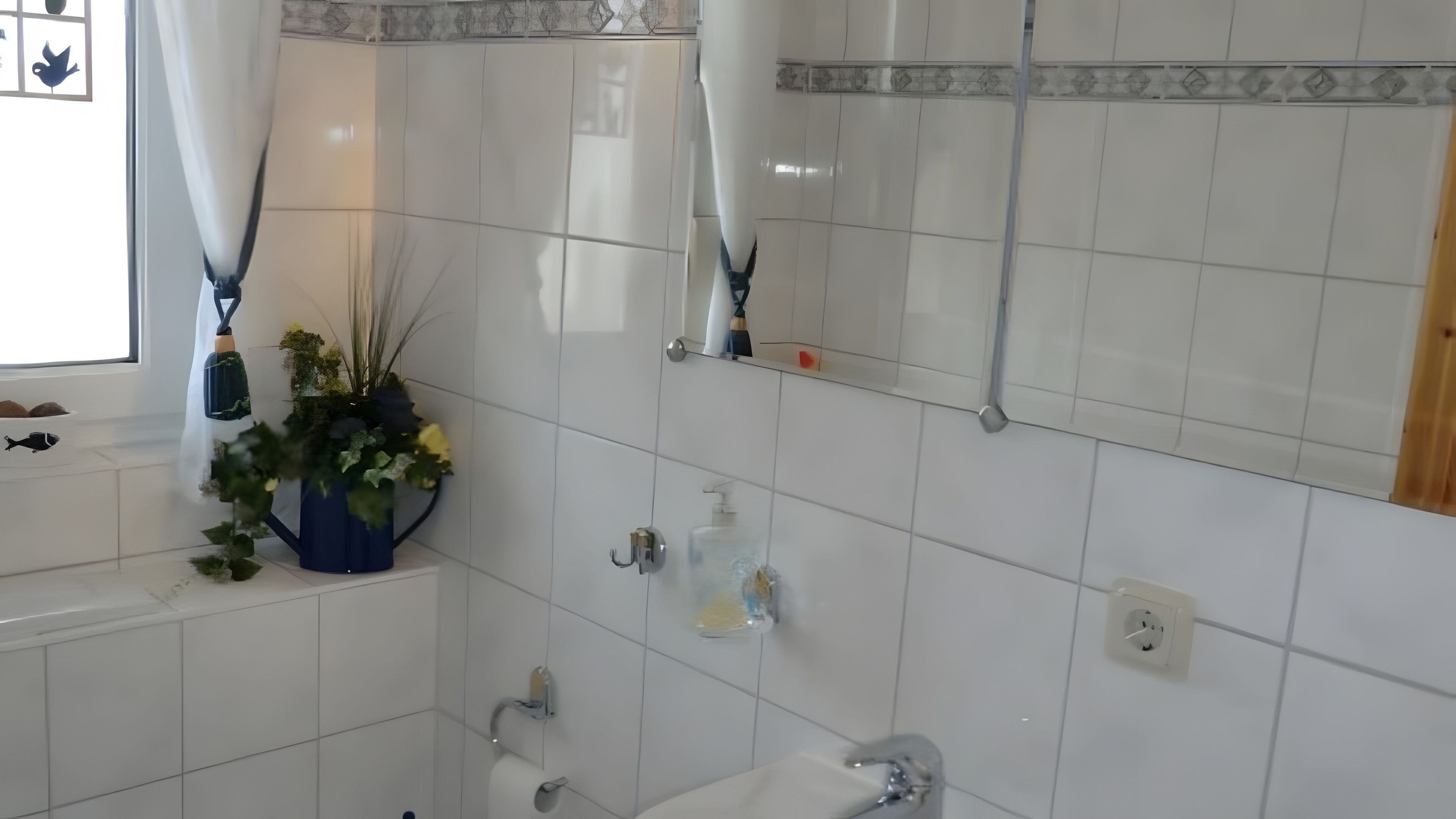 Une salle de bain avec des carreaux blancs, un lavabo, un miroir et une fenêtre avec des rideaux.