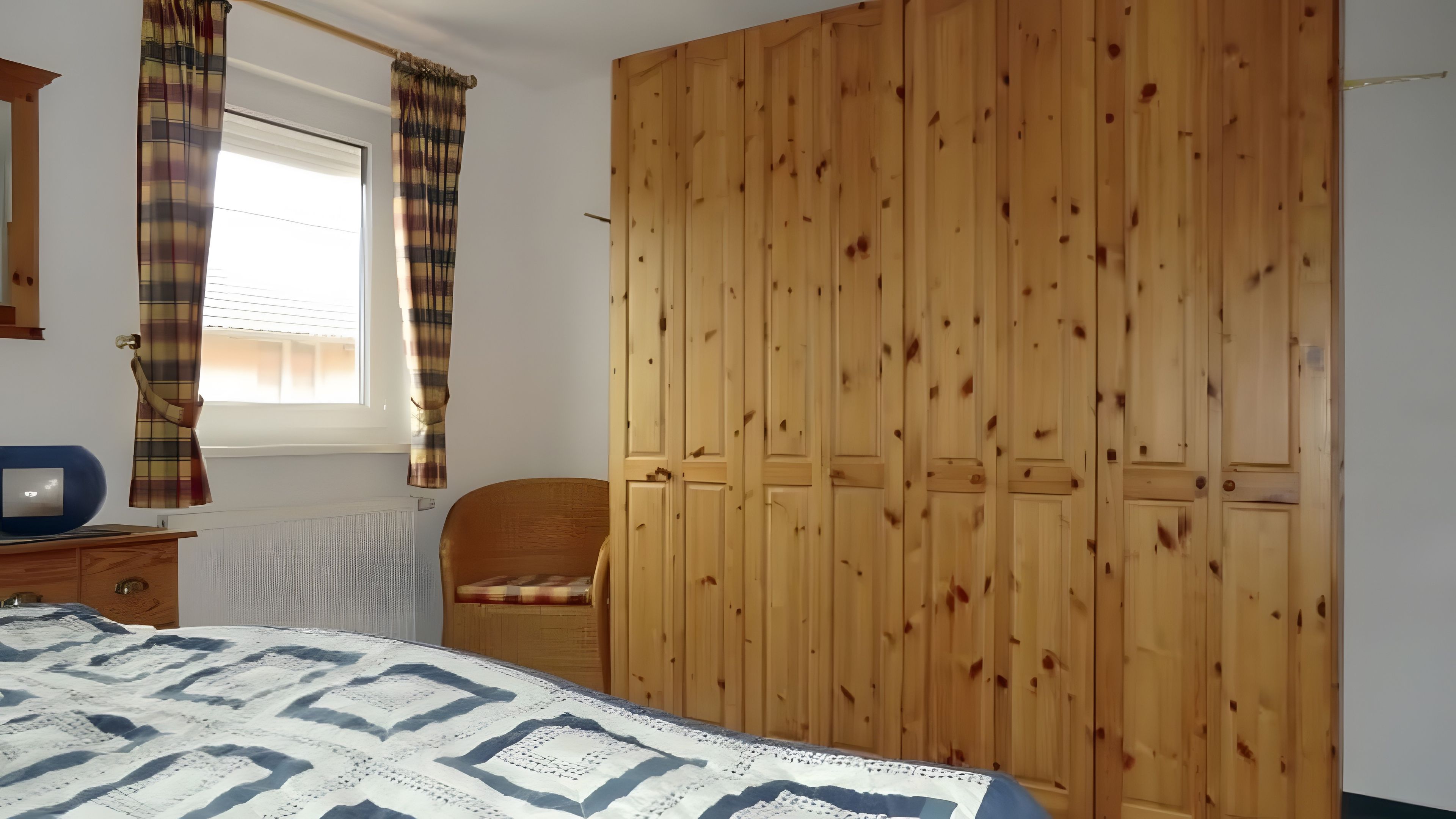 A bedroom with a bed, a wooden wardrobe, a chair, and a window with curtains.