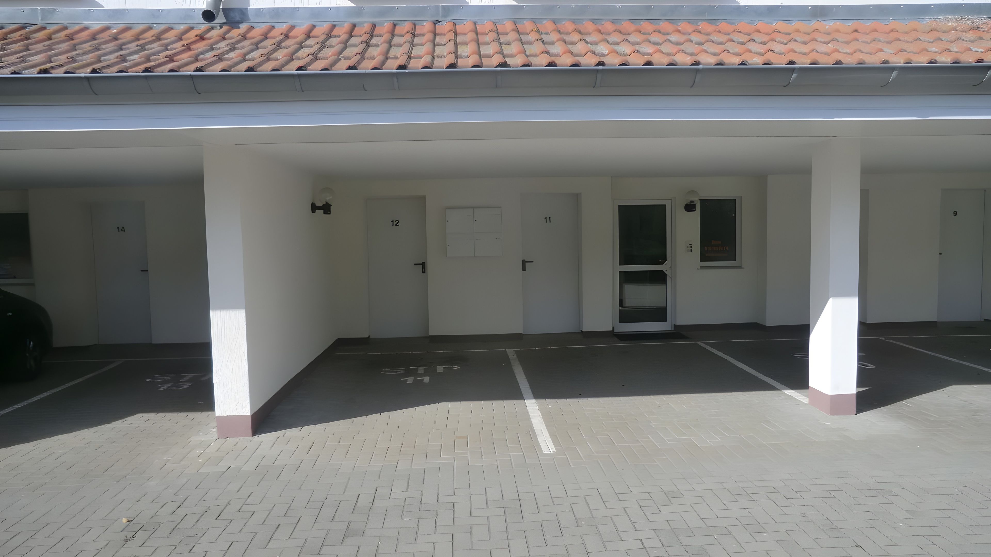 Un espace de stationnement couvert, doté de portes numérotées, d’un toit en tuiles et d’un sol pavé de briques.