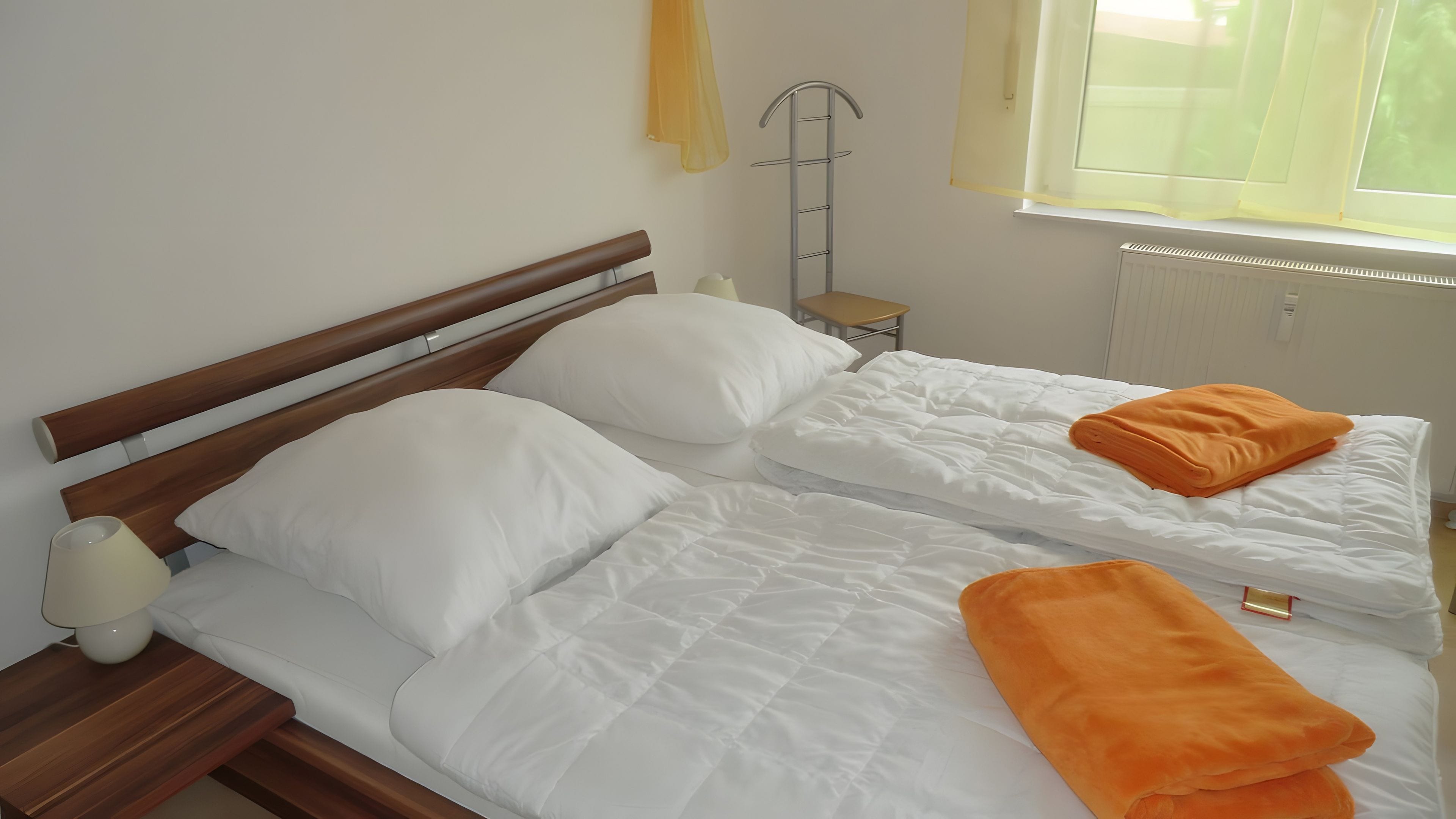 Ein Doppelbett mit weißer Bettwäsche und einem orangefarbenen Handtuch, ein Nachttisch mit Lampe sowie ein Fenster mit Jalousien.