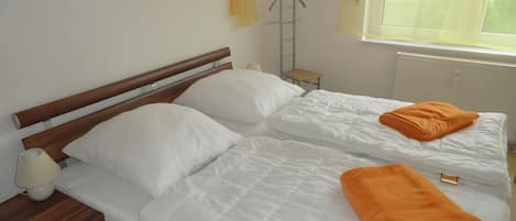 Ein Doppelbett mit weißer Bettwäsche und einem orangefarbenen Handtuch, ein Nachttisch mit Lampe sowie ein Fenster mit Jalousien.