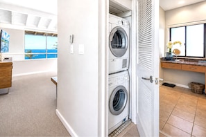 A laundry room with stacked washers and dryers, a wooden bench, and a view of the ocean through a window. - Kapalua Bay Villas KBV-32B3 - Ocean View 1 BR 180 Degree Views - Steps to Beach (Lahaina)