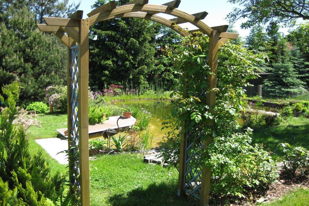 Eine Holz-Pergola in einem Garten mit Teich, umgeben von üppiger Bepflanzung und Bäumen.