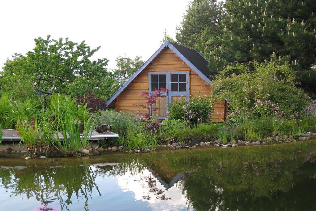 A wooden cabin with a pond in front, surrounded by lush greenery and flowering plants.