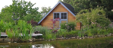 A wooden cabin with a pond in front, surrounded by lush greenery and flowering plants.