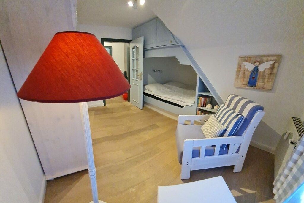 A room with a bunk bed, a white chair with striped cushions, a red floor lamp, and a wall-mounted shelf with books.