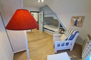 Een kamer met een stapelbed, een witte stoel met gestreepte kussens, een rode vloerlamp en een wandplank met boeken.