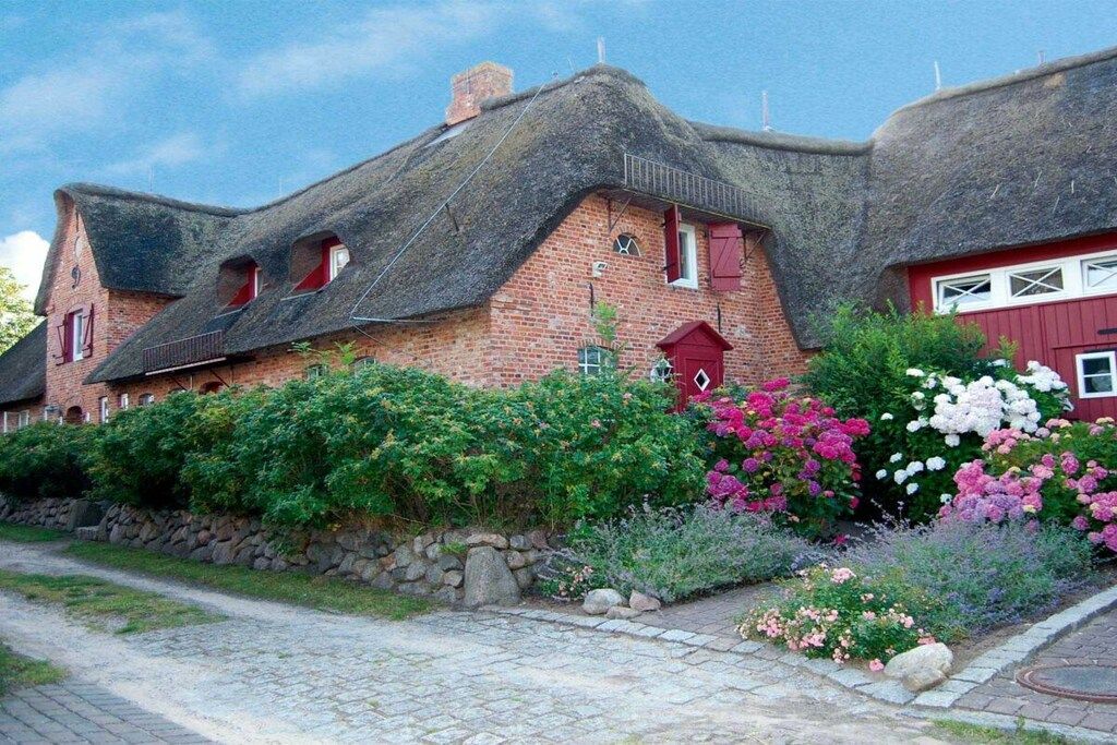 A traditional brick house with a thatched roof, a stone wall, and a garden with various flowers.