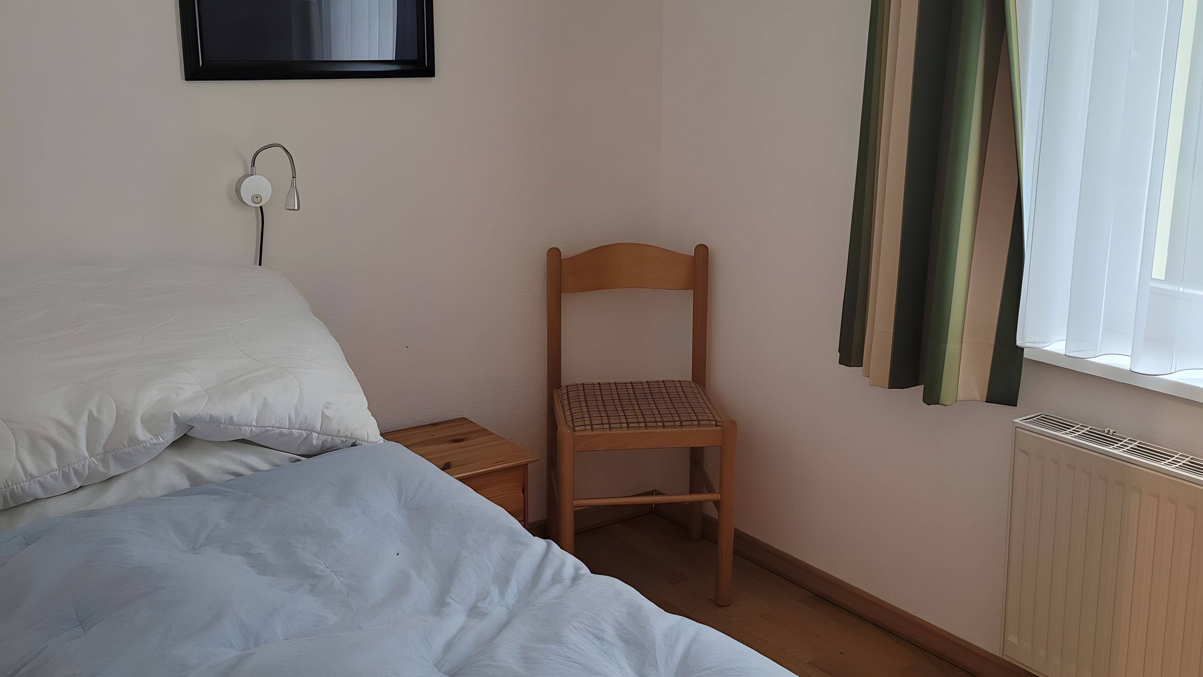 Een slaapkamer met een bed, een houten stoel, een nachtkastje, een stopcontact op de muur en een raam met gordijnen.