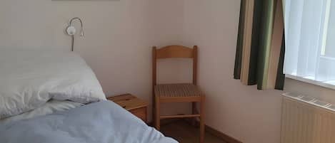 Quarto com cama, cadeira de madeira, mesa de cabeceira, tomada na parede e janela com cortinas.