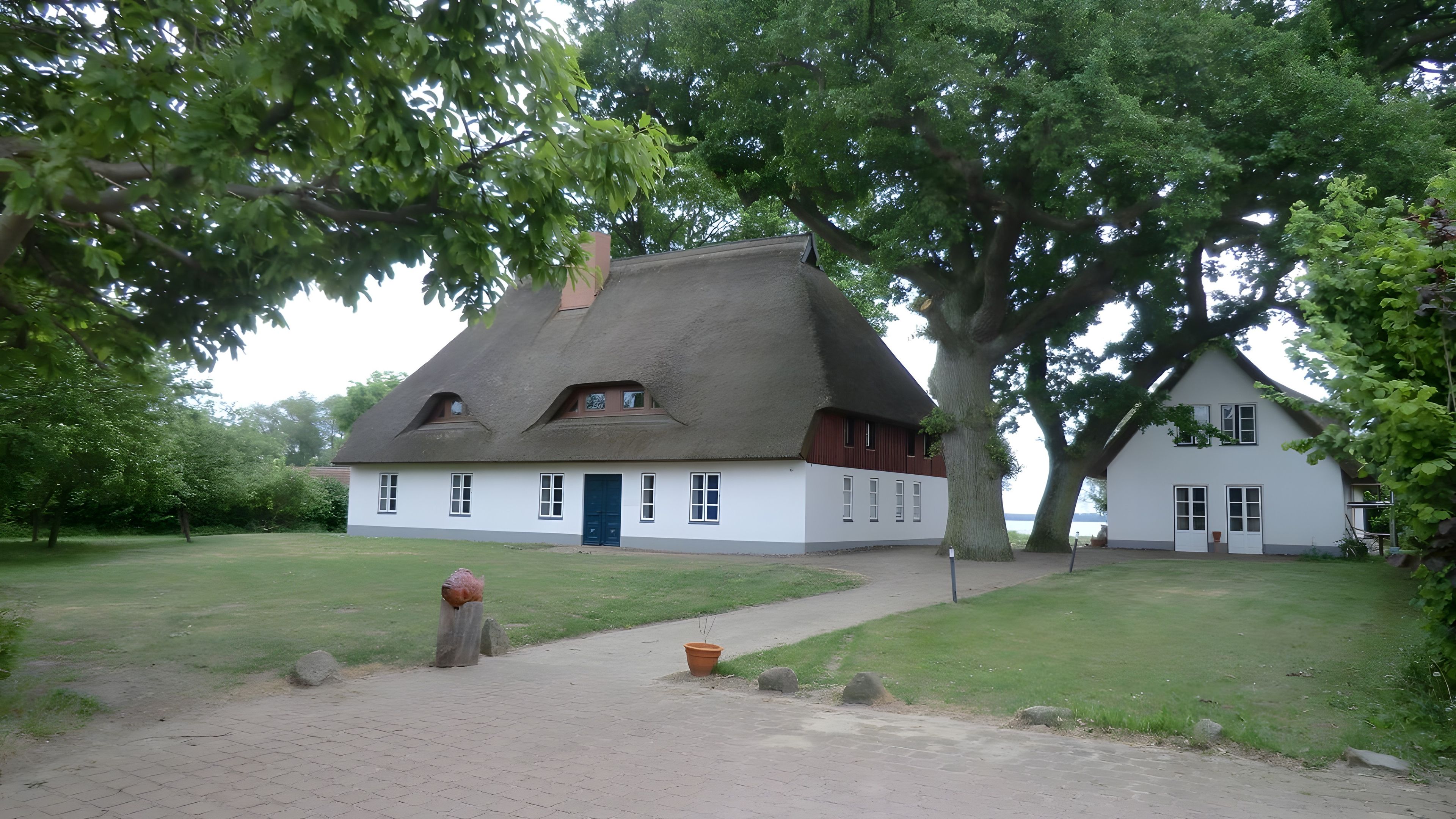 Une maison traditionnelle avec un toit de chaume, entourée d’une pelouse bien entretenue et de grands arbres.