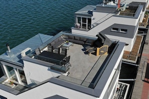 Eine moderne Dachterrasse mit Sitzgelegenheit, einem kleinen Tisch und einem Glasgeländer mit Blick auf das Wasser.