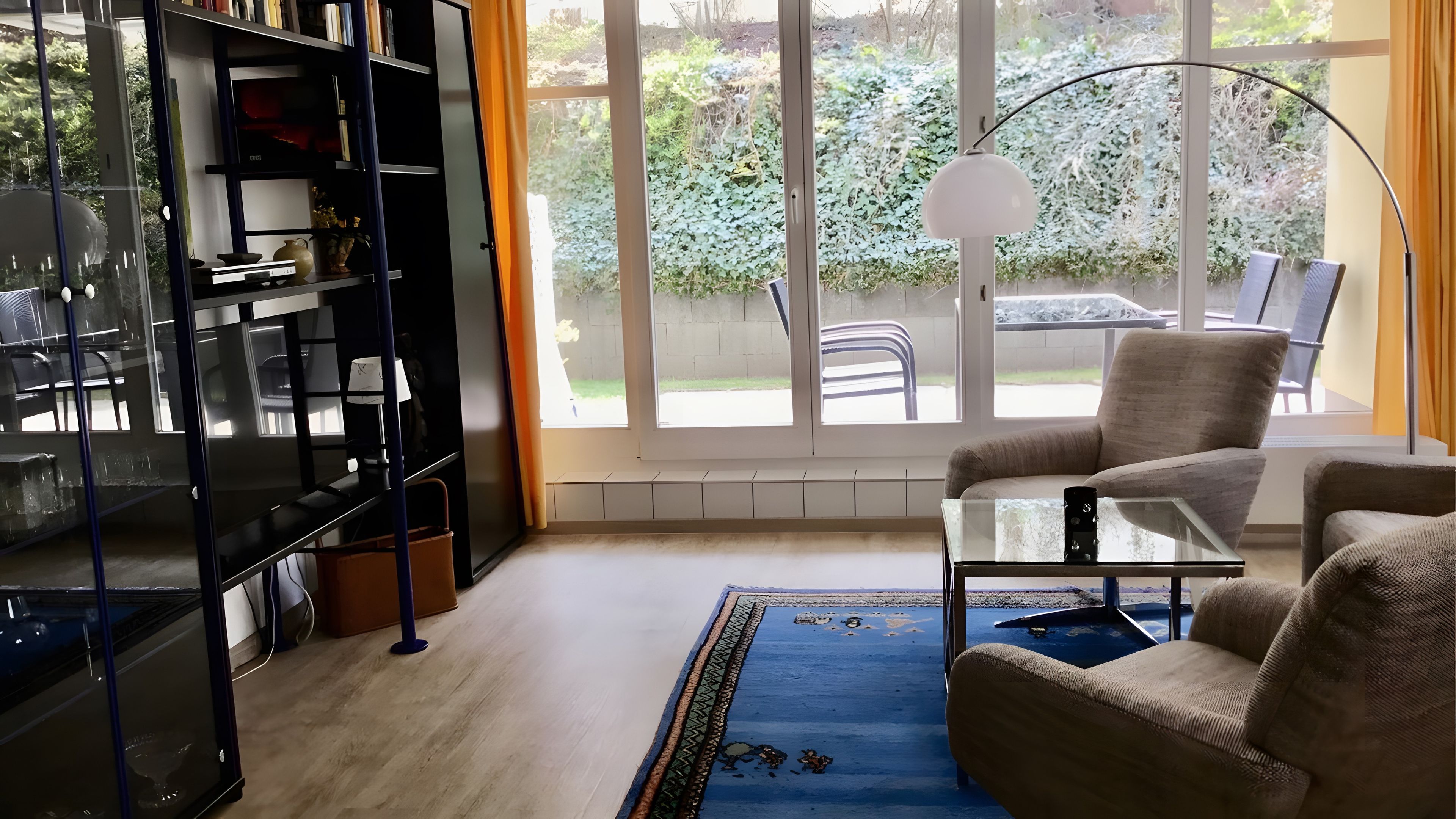 Ein Zimmer mit Bücherregal, Glastisch, Sofa, einem gemusterten Teppich und Blick auf einen Garten durch große Fenster.