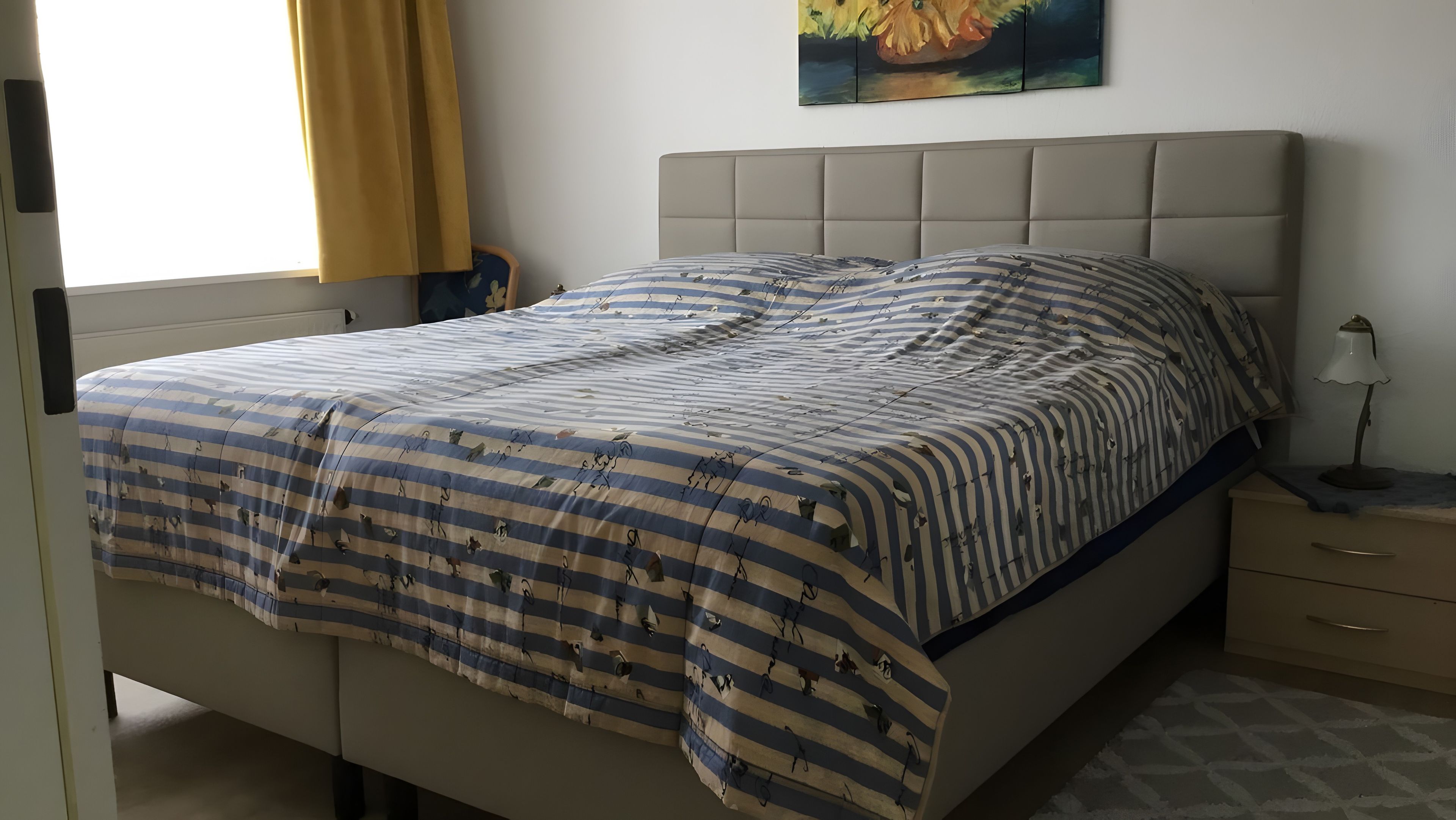 Ein Bett mit gestreifter Bettdecke, ein Kopfteil, ein Nachttisch mit Lampe und ein Bild an der Wand.