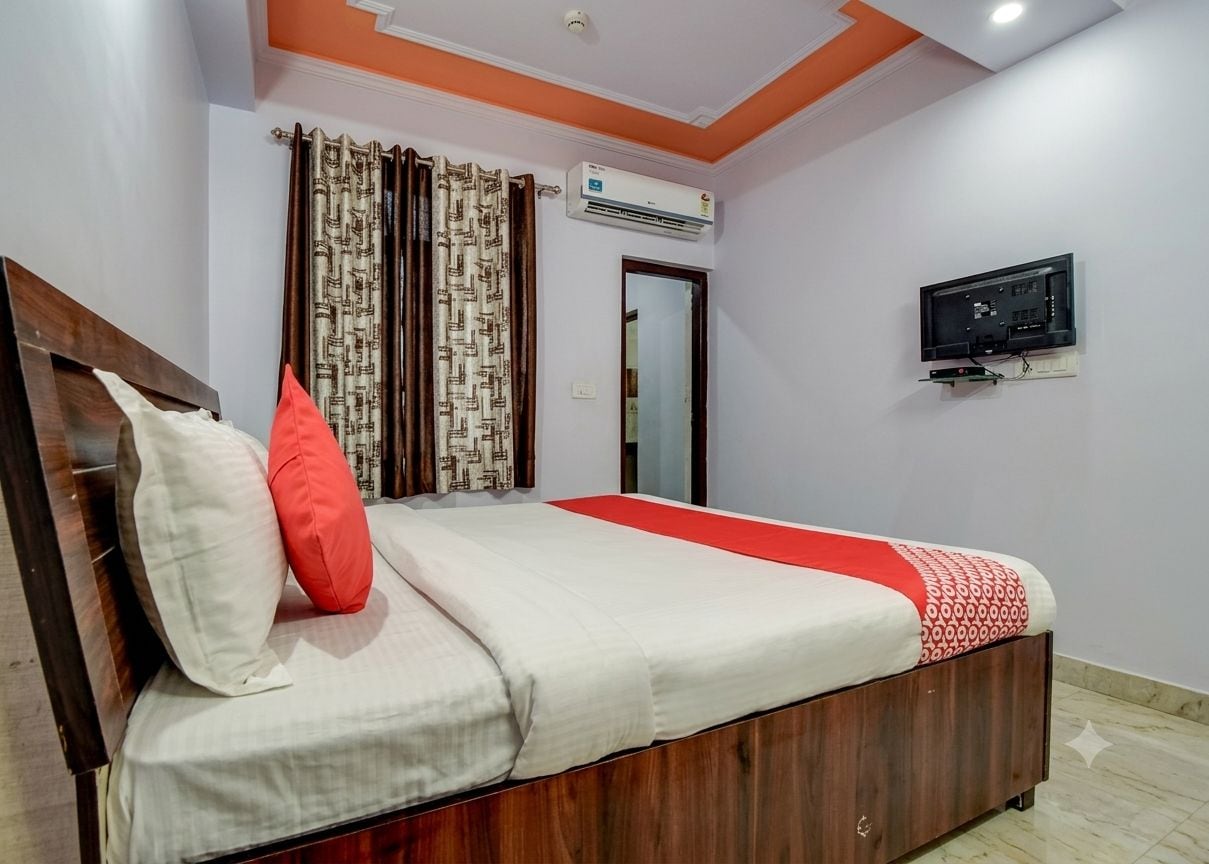 A bedroom with a bed, curtains, an air conditioning unit, a television, and a door.