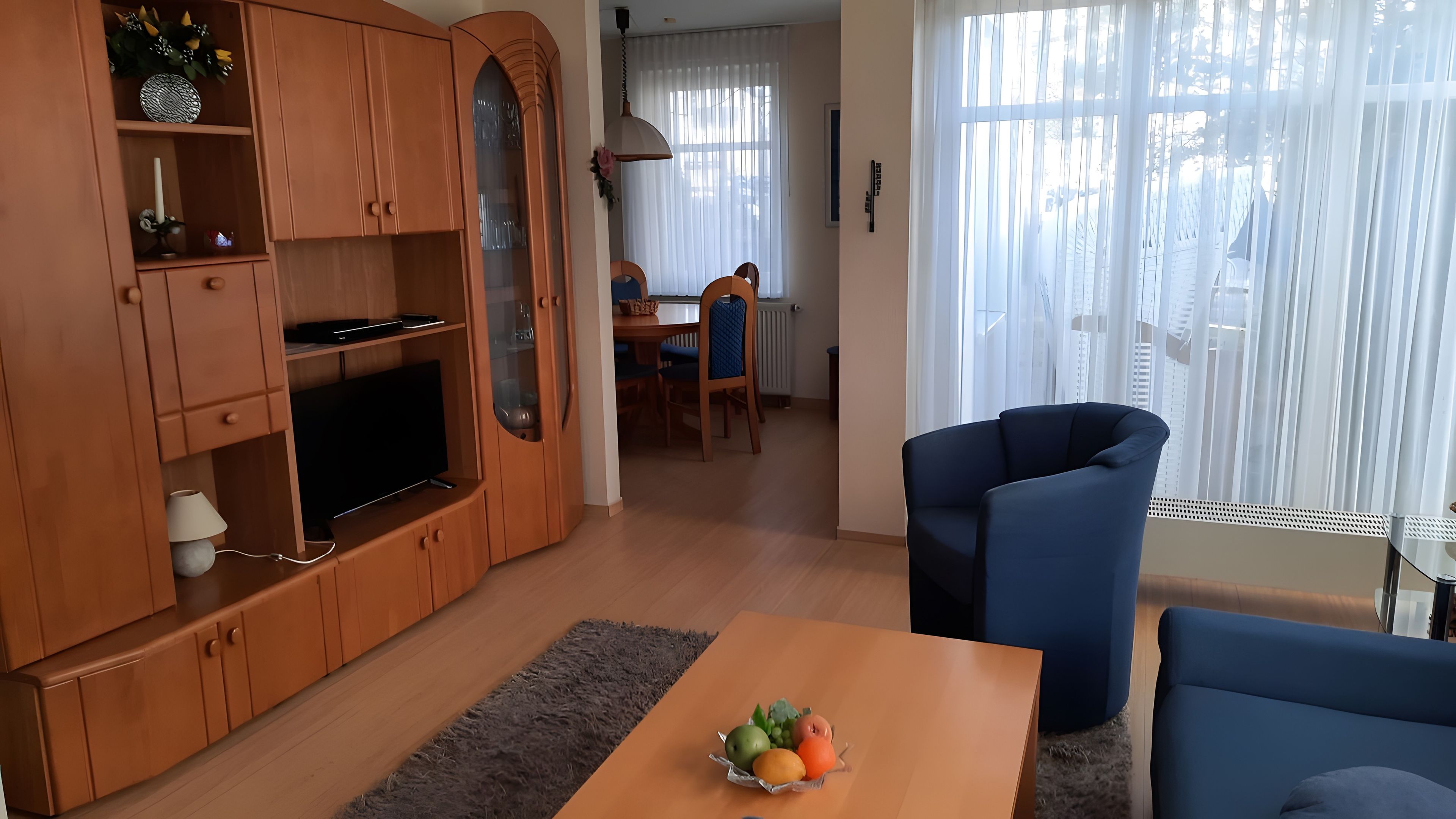 Ein Wohnzimmer mit Fernseher, Holzschränken, einem Esstisch mit Stühlen und einem blauen Sessel.