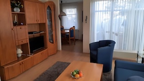 Ein Wohnzimmer mit Fernseher, Holzschränken, einem Essbereich mit Tisch und Stühlen sowie einem großen Fenster mit Vorhängen.