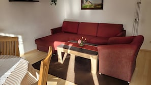 Ein rotes Sofa, ein Holzstuhl, ein kleiner Tisch mit Blumenvase und ein Gemälde an der Wand.
