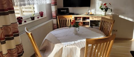Zona pranzo con tavolo rotondo, sedie, televisione e finestra con tende.