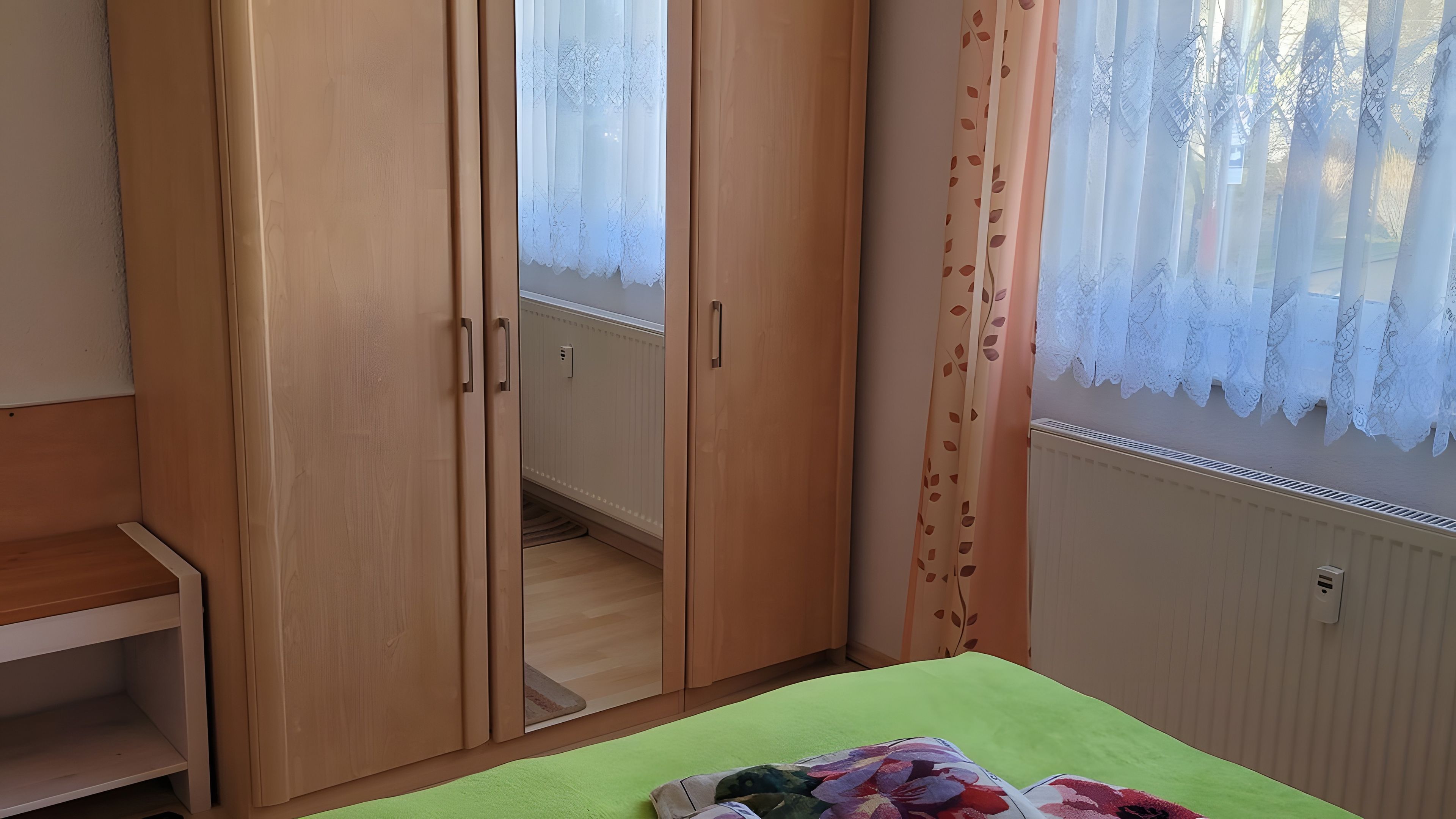 Ein Zimmer mit Kleiderschrank, einem Bett mit grüner Bettdecke und einem Fenster mit Spitzenvorhängen.