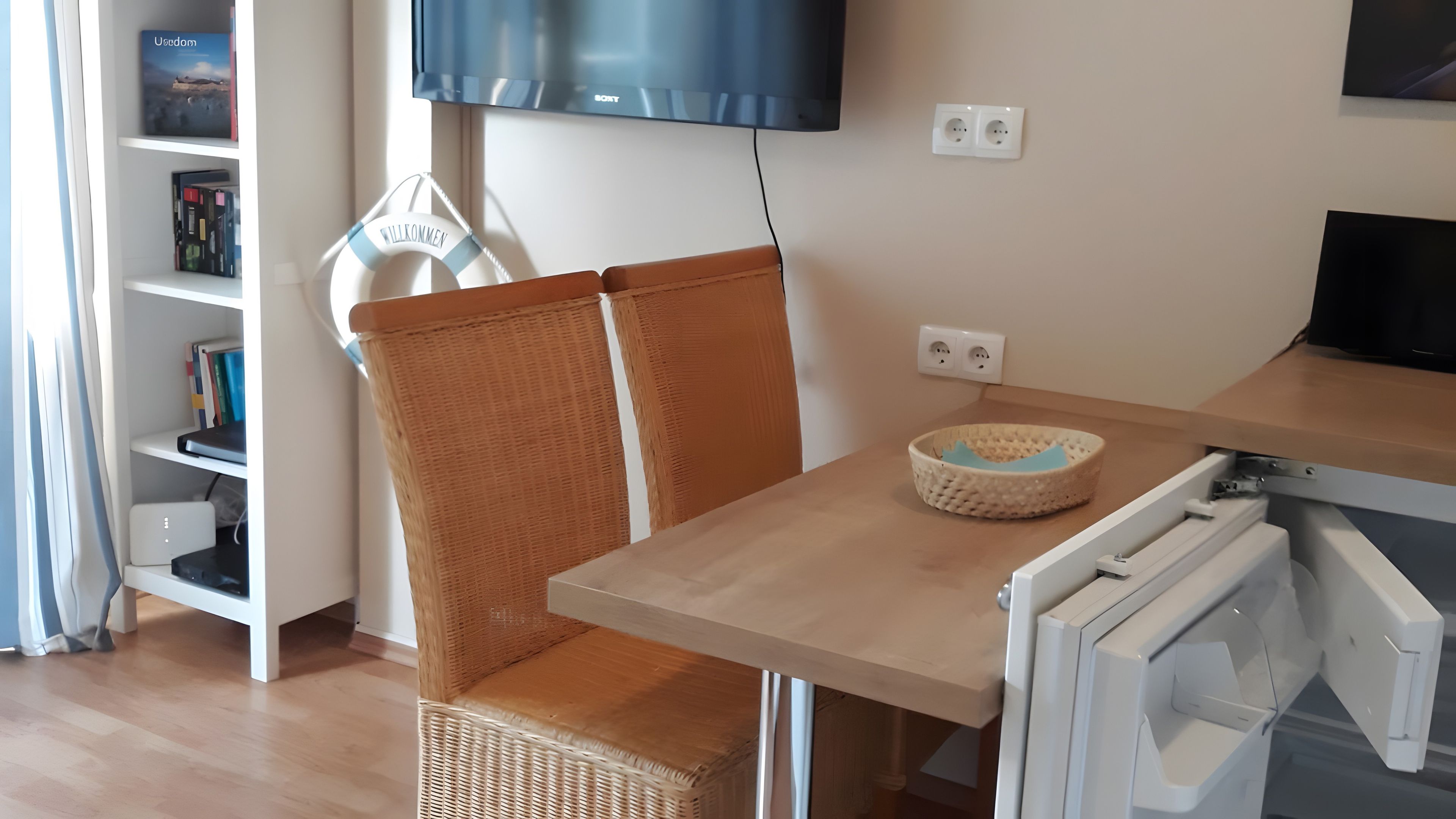 Ein Zimmer mit einem Korbsessel, einem Tisch, einem Flachbildfernseher, einem Bücherregal und einer Steckdose.