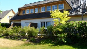 Une maison à deux étages, dotée d’un toit en pente, de nombreuses fenêtres et d’un jardin où poussent diverses plantes et fleurs.