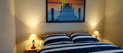 Habitación de hotel con una cama, dos lámparas de noche, un edredón a rayas y un cuadro de un atardecer en la pared.