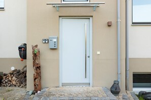 Eine beige farbene Außenwand mit einer weißen Tür, einem Briefkasten und einer kleinen Mülltonne.