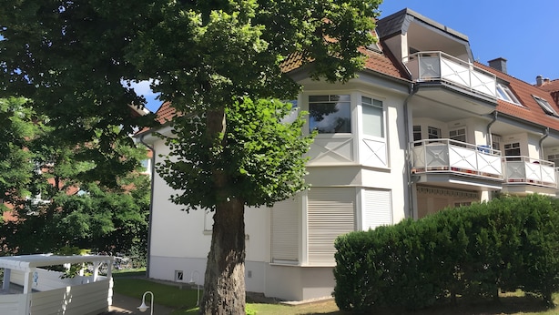 Un immeuble résidentiel de deux étages doté d’un balcon, avec un arbre au premier plan et un ciel dégagé.