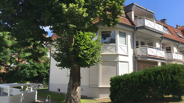Ein zweistöckiges Wohngebäude mit Balkon, ein Baum im Vordergrund und ein klarer Himmel.