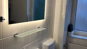 Ein Badezimmer mit weiß geflieste Wand, einem Spiegel, einem Ablagefach und einem Fenster.