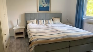 Une chambre à coucher avec un lit, une table de chevet, une lampe, un tableau encadré au mur et une fenêtre avec des rideaux.