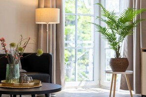 Ein Zimmer mit einem runden Tisch, einer Couch, einer Lampe, einem Fenster mit Vorhängen und einer Topfpflanze.