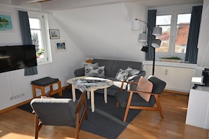 Ein modernes Wohnzimmer mit grauem Sofa, Couchtisch und Fernseher. Zwei Fenster mit Vorhängen, eine Lampe und eine kleine Pflanze auf der Fensterbank.