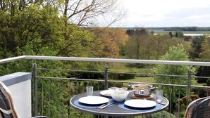 A balcony with a table set for two, overlooking a scenic landscape with trees and a distant view of a body of water.