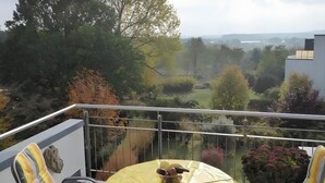 Un balcó amb taula i cadires, amb vista a un jardí i un paisatge llunyà.