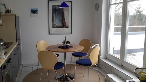 A modern kitchen with a dining area, a potted plant on the table, and a view through the window.