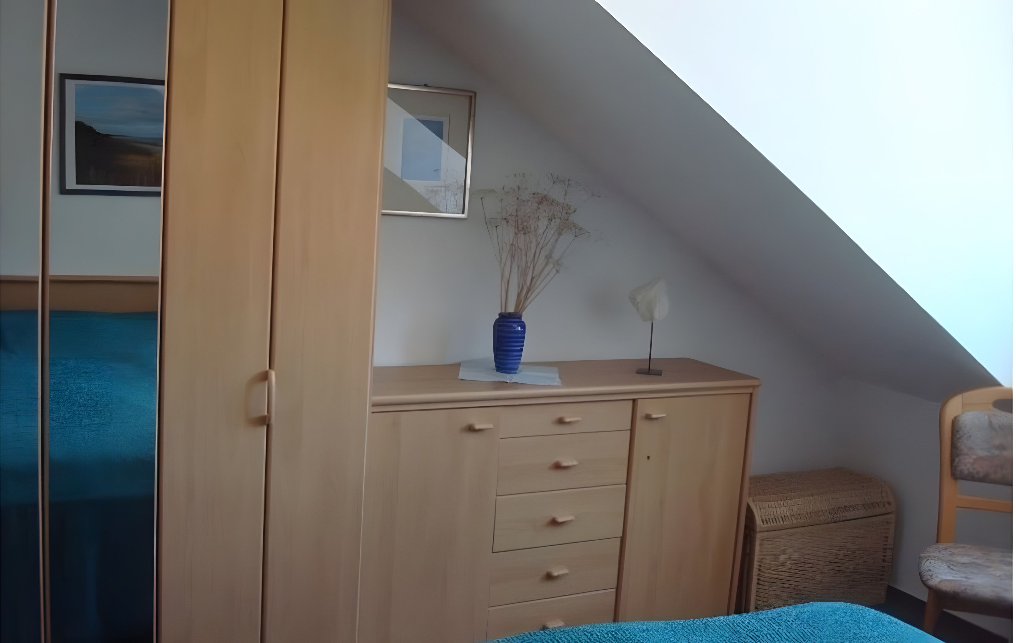 Ein Zimmer mit schrägem Dach, einem Holzschrank mit Schubladen, einem kleinen Korbstuhl-Beistelltisch und einem gerahmten Bild an der Wand.